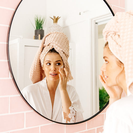 Image of young woman in a bathrobe, looking at herself in a round mirror, putting on a face mask. natural australian clay mask